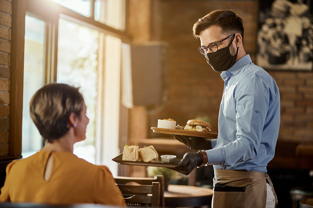 Server at restaurant with face mask serving woman sitting down