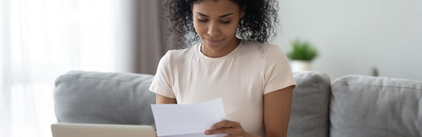 Woman sitting on couch with laptop in front of her looking at a letter she received in the mail