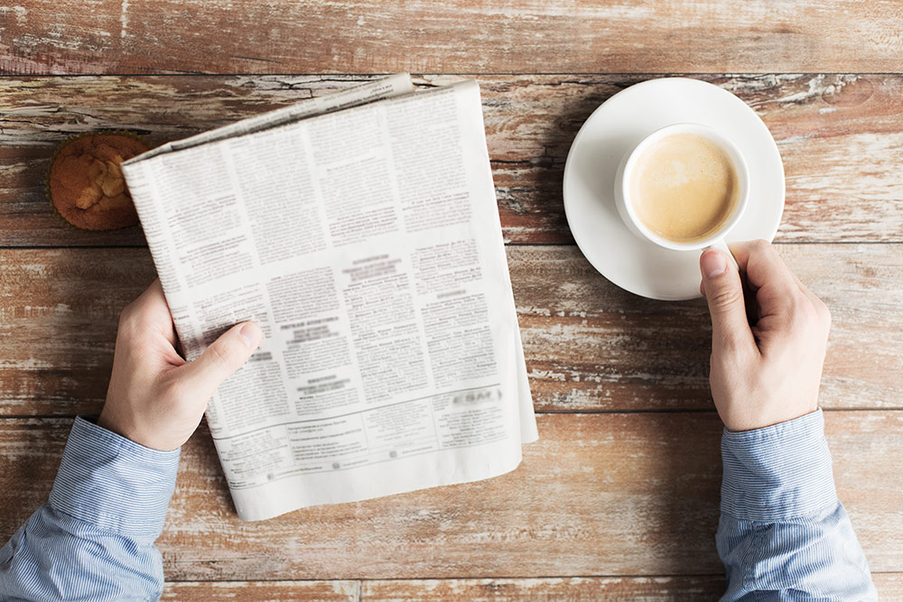 Close up of person reading newspaper at table with hand on coffee mug