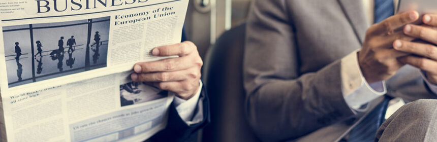 Two men sitting on a bench, one with a newspaper the other on their smartphone