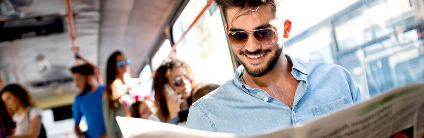 Man sitting on a bus smiling and wearing sunglasses while reading the newspaper