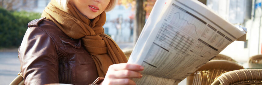 A young woman reading a newspaper while sitting outdoors at a cafe