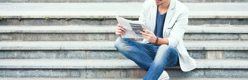 A young man dressed in business casual sits reading a newspaper on a flight of steps
